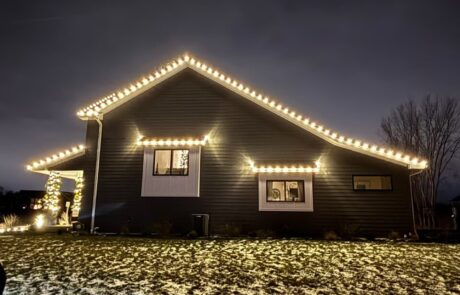 Warm-white roofline lights tracing the gables of a home with a lit garland arch, installed by Lighting Squad