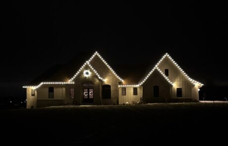 Warm-white roofline lights outlining the peaks of a stone-and-siding home with a lit wreath, by Lighting Squad